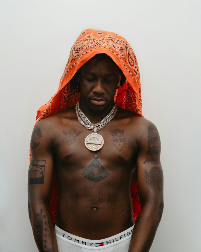 Tattooed man with orange bandana, adorned with jewelry in an introspective pose.